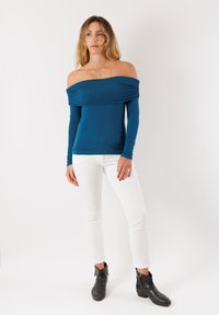 Top teal senza spalle con maniche lunghe, design arricciato sul colletto. Abbinato a pantaloni bianchi a taglio slim e stivali neri alla caviglia.