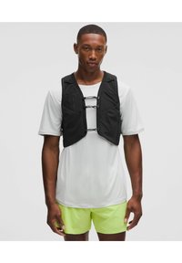 Chaleco de entrenamiento negro con un diseño ajustado, que presenta cremalleras y correas elásticas, usado sobre una camiseta gris claro y pantalones cortos amarillos.