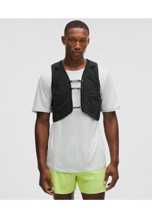 Svart träningsväst med en figurnära design, med blixtlås och elastiska remmar, bärs över en ljusgrå skjorta och gula shorts.