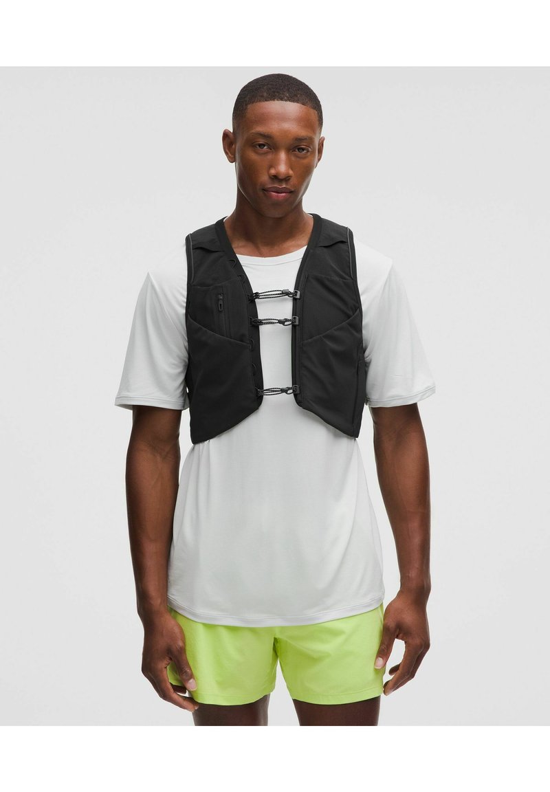 Mustat koulutusliivit, joissa on vartalonmyötäinen muotoilu, jossa on vetoketjuja ja joustavia hihnoja, käytetään vaaleanharmaan paidan ja keltaisten shortsien päällä.