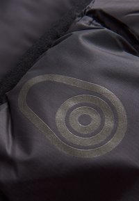 Svart tyg med en subtil rutmönster har en cirkulär logotyp i reflektionsgrå som är placerad nära en mjuk, texturerad kant.