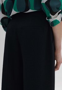Pantalons larges texturés noirs avec un détail de poche arrière, associés à un blouse à motifs verts et noirs.