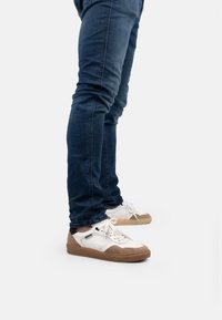 Zapatillas de cuero blanco con detalles de ante en beige y suelas de goma marrón claro, combinadas con unos vaqueros ajustados de color azul oscuro. Los zapatos cuentan con un diseño perforado.