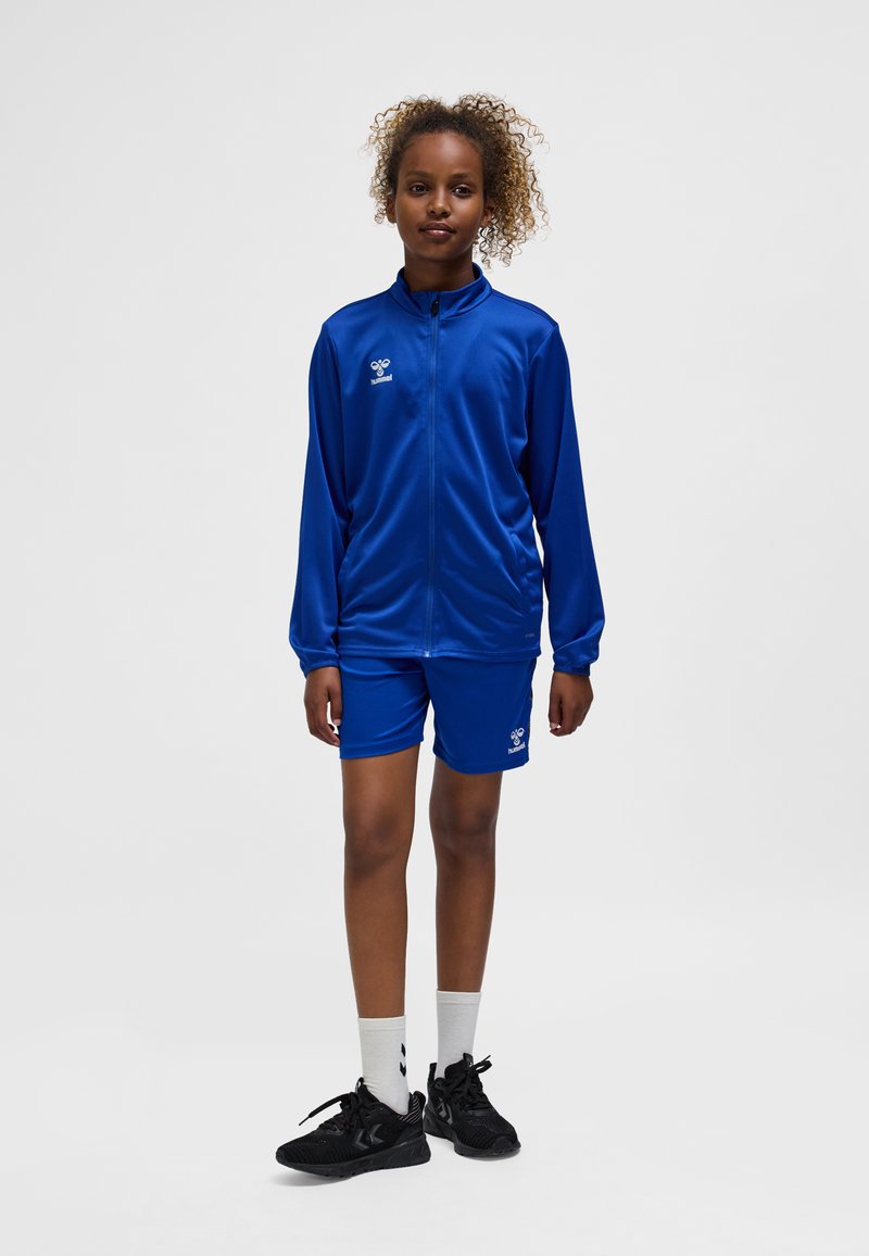 Blauer Sportanzug aus glattem, glänzendem Stoff, bestehend aus einer Jacke mit Reißverschluss und passenden Shorts, kombiniert mit schwarzen Sportschuhen und weißen Socken.