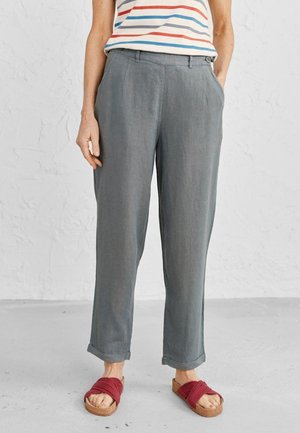 Pantaloni in lino grigi con una vestibilità comoda, dotati di tasche frontali e orli arrotolati, abbinati a sandali rossi intrecciati su un pavimento texturizzato.