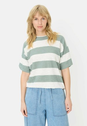 Kvinde med blondt hår iført en løstsiddende grøn og hvid stribet kortærmet skjorte og lyseblå højttaljede denimbukser.