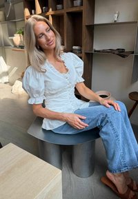 Femme aux cheveux blonds portant un chemisier blanc et un jean bleu, assise sur un tabouret métallique moderne dans une pièce lumineuse avec des étagères et de la poterie.