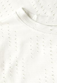 Tessuto di cotone bianco con un motivo testurizzato caratterizzato da piccoli fori distribuiti uniformemente, scollo rotondo e bordo a costine sul colletto.