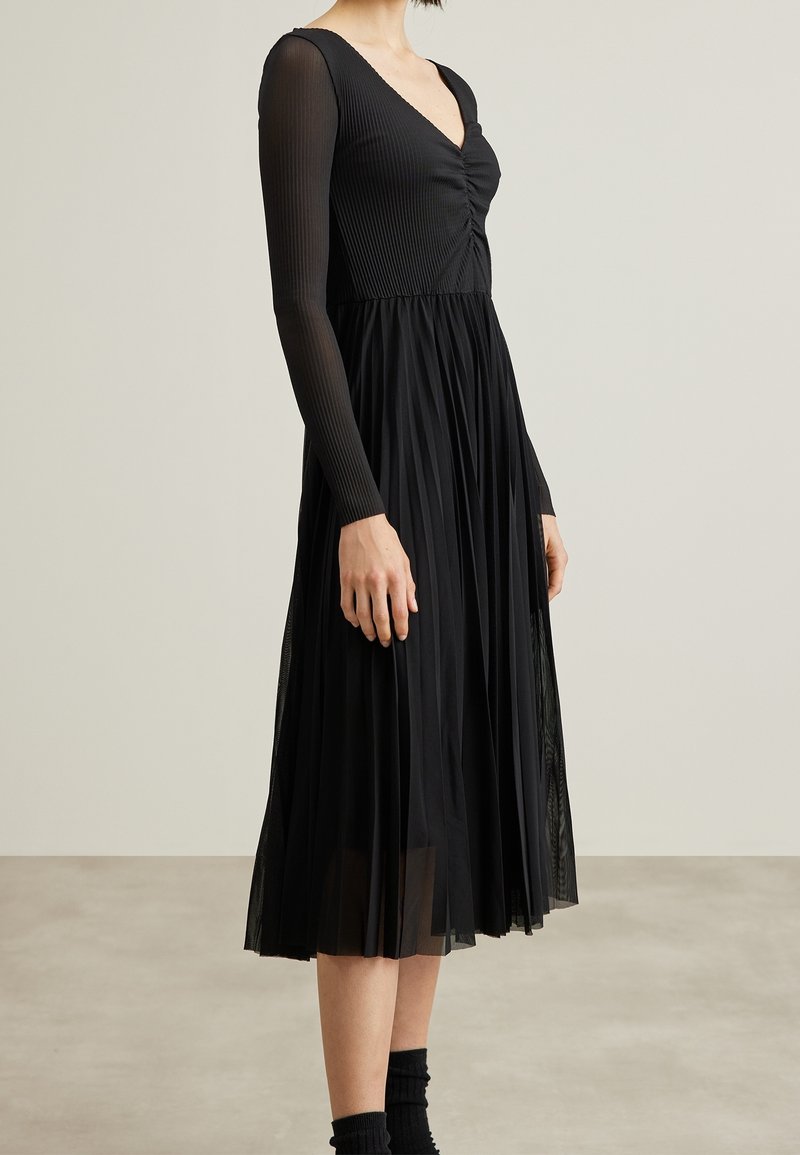 Femme portant une robe midi noire plissée à manches longues avec un col en V et un détail froncé sur la poitrine, debout devant un fond uni.
