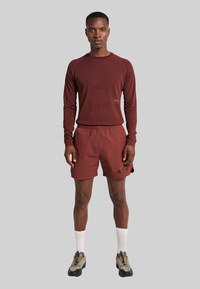 Homme portant un t-shirt à manches longues bordeaux, des shorts assortis avec le texte noir "ATW", des chaussettes blanches et des chaussures de course grises, debout devant un fond gris.