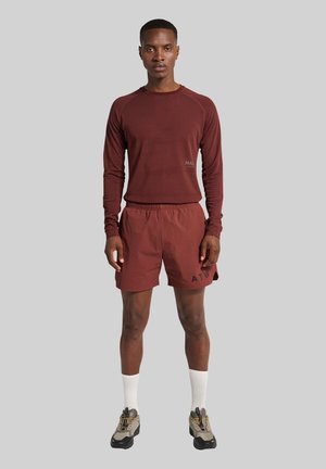Homme portant un t-shirt à manches longues bordeaux, des shorts assortis avec le texte noir "ATW", des chaussettes blanches et des chaussures de course grises, debout devant un fond gris.