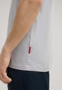Lichtgrijs katoenen T-shirt met ronde hals, korte mouwen, zijsplitten en een klein rood label aan de zoom. Glad textuur, casual pasvorm.