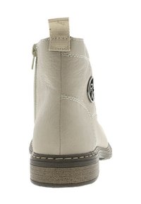 Botte de cheville beige en matériau synthétique texturé, avec une fermeture éclair latérale, un tirant en tissu et un logo circulaire à l'arrière près d'une semelle en bois cousue.