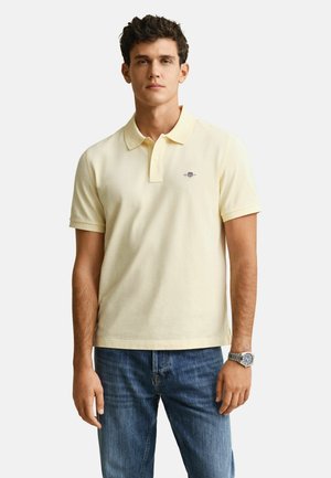 Junger Mann, der ein hellgelbes Polohemd mit kurzen Ärmeln, blaue Jeans und eine silberne Armbanduhr am linken Handgelenk trägt.