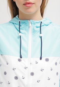 Jasnoniebieska kurtka z kapturem, dolna część w kolorze białym z wzorami granatowych kotwic i kół sterowych. Wyposażona w sznurki ściągające i zamek błyskawiczny.