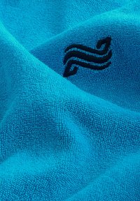 Blå badhandduk i frotté med en mjuk, texturerad yta; har en broderad svart virvel-logotyp.