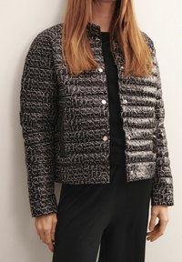 Schwarze gesteppte Jacke mit einem weißen geometrischen Muster, Druckknöpfen und einem verkürzten Design. Tragen über einem schwarzen Oberteil und schwarzen Hosen.