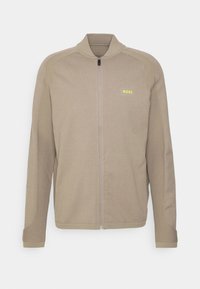 Světle hnědá zipová bunda vyrobená z texturované tkaniny, s vysokým límcem, dlouhými rukávy a malým žlutým logem "BOSS" na hrudi.