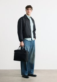 Νέος άνδρας με μαύρο σακάκι, ανοιχτό μπλε πουκάμισο και τζιν παντελόνια φαρδιάς γραμμής, κρατώντας μια μαύρη τσάντα, στέκεται μπροστά σε λευκό τοίχο σε εσωτερικό χώρο.