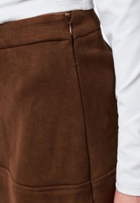 Nahaufnahme einer Person, die einen braunen Wildlederrock mit seitlichem Reißverschluss trägt, kombiniert mit einem weißen Langarmoberteil.