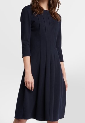 Femme portant une robe bleu marine arrivant aux genoux, avec des manches trois-quarts et des plis verticaux sur le devant, sur un fond uni.