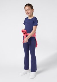 Chica de pie y sonriendo, vestida con una camiseta de Champion azul marino y pantalones, con una sudadera rosa atada alrededor de la cintura y zapatillas blancas.