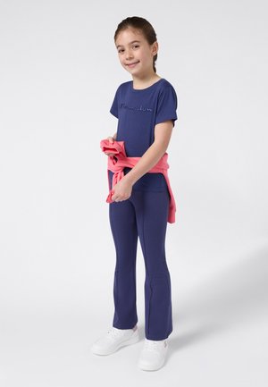 Meisje staat rechtop en glimlacht, draagt een marineblauw Champion shirt en broek met een roze sweatshirt om haar middel geknoopt, witte sneakers.