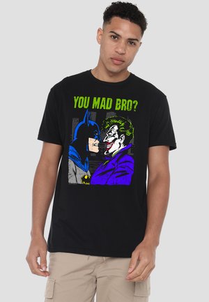 Young man wearing a black t-shirt showing Batman and Joker facing each other with text "YOU MAD BRO?" in green above.