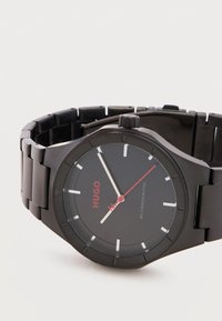 Μαύρο μεταλλικό ρολόι χειρός με στρογγυλό καντράν, κόκκινη δείκτη δευτερολέπτου, λευκούς δείκτες ώρας/λεπτών και τη λέξη «HUGO» με κόκκινα γράμματα στο καντράν.