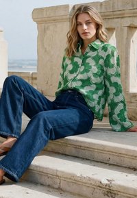 Mladá žena sediaca na kamenných schodoch, oblečená v zelenej kvetinovej blúzke, širokých modrých džínsoch a sandáloch, s blond vlasmi a neutrálnym výrazom tváre.