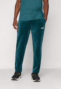 Turkusowe welurowe dresy o slim fit i subtelnym połysku, z białym logo Puma na lewym udzie. W połączeniu z czarnymi butami sportowymi.