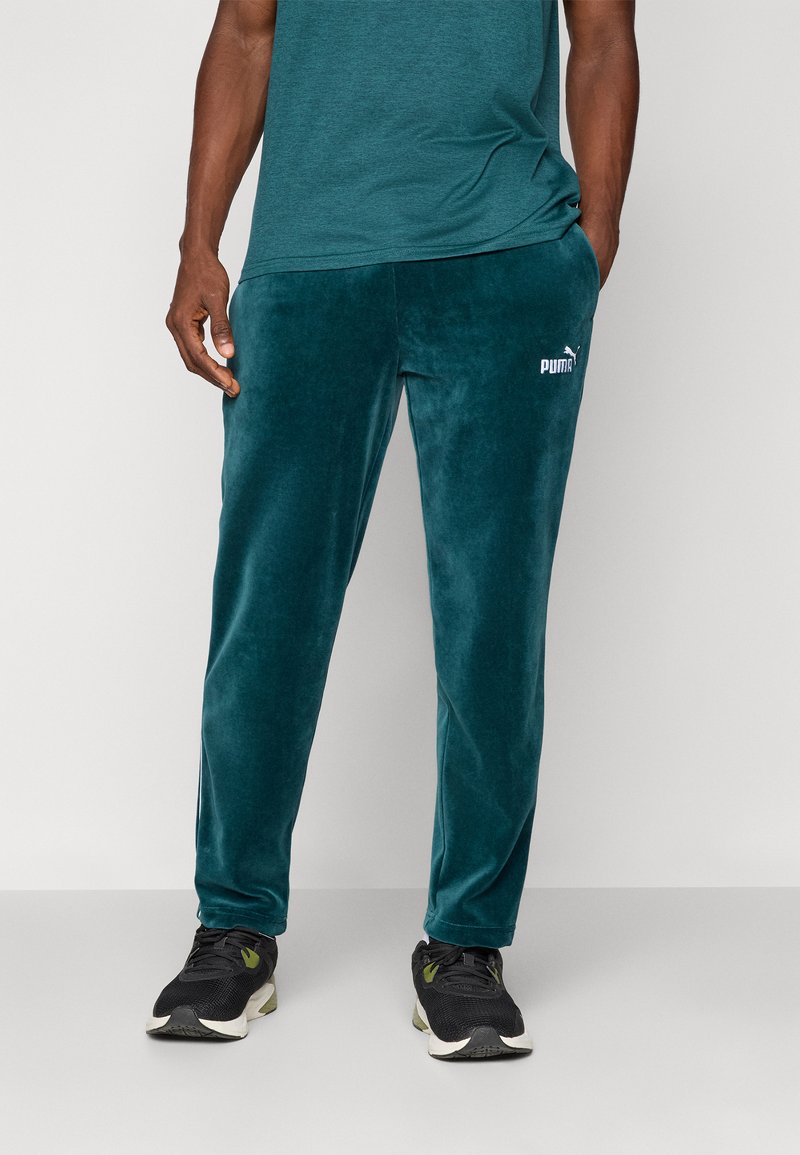 Turkusowe welurowe dresy o slim fit i subtelnym połysku, z białym logo Puma na lewym udzie. W połączeniu z czarnymi butami sportowymi.