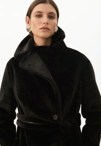 Černý kabát z umělé kožešiny s velkým límcem, dlouhými rukávy a páskem v pase. Obsahuje jedno knoflíkové zapínání a jemnou texturu.