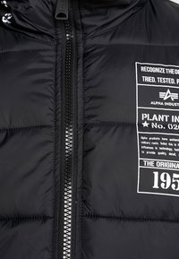 Giacca nera imbottita con zip frontale e etichetta stampata bianca che riporta i dettagli "Alpha Industries" e "Plant No. 020".