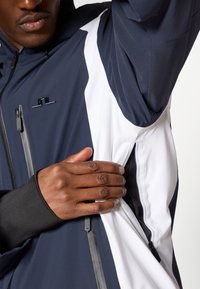 Tengerész-kék és fehér dzseki könnyű, vízálló kialakítással rendelkezik, cipzáras oldalzsebbel és eltérő textúrákkal. Kis logóval a mellkason.