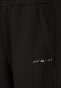 Zwarte sportbroek met een elastische tailleband en zijzakken. Kenmerkt zich door een zilveren "ENDURANCE" logo op het onderbeen. Zachte, gladde stoftextuur.