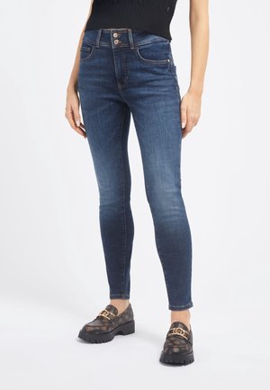 Frau trägt dunkelblaue Skinny Jeans mit Doppelknopf-Bund und schwarzen gemusterten Loafers mit goldener Kettendetail.