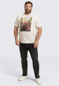 Lichtcrème graphic t-shirt met een auto foto ontwerp, gecombineerd met zwarte jeans en witte sneakers, staand tegen een neutrale achtergrond.