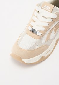 Sneaker beige con un mix di materiali in suede e rete, lacci bianchi, logo argentato e una suola bianca imbottita.