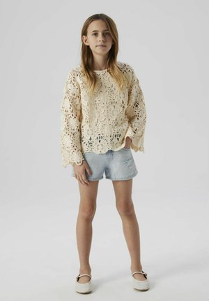Top de ganchillo en blanco roto, con patrones florales y mangas acampanadas, combinado con pantalones cortos de mezclilla desgastados y zapatos de color claro.
