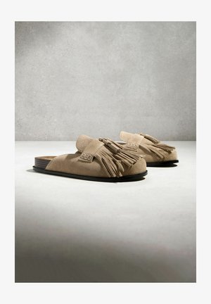 Beżowe zamszowe buty wsuwane z frędzlami na górze, czarne płaskie podeszwy i odkryte zapiętki, umieszczone na białej powierzchni na szarym tle.