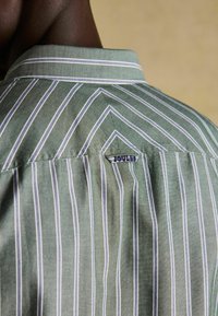 Grün gestreiftes Hemd mit weißen und violetten Streifen, ausgestattet mit einem spitzen Kragen und einem kleinen blauen Etikett auf der Rückseite. Die Stofftextur wirkt glatt.