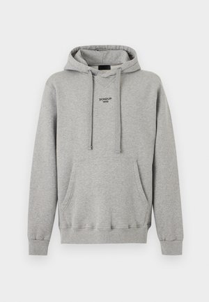 Jeune homme debout, portant un sweat à capuche gris avec poche avant, un jean bleu et des baskets noires avec semelles blanches, sur un fond blanc uni.