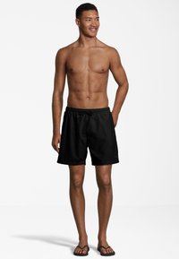 Costumi da bagno neri con vita elastica, coulisse regolabile e texture liscia. Indossati con ciabatte nere. Branding minimale sui costumi.