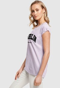 Lyslilla t-shirt med cap-sleeve og sort "BERLIN" tekst. Fremstillet af bomuld med en blød tekstur og afslappet pasform, med trimmet ved ærmerne.
