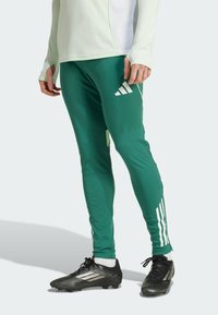 Zielone spodnie sportowe z białymi paskami, wykonane z lekkiego materiału. Charakteryzują się dopasowanym krojem i logo Adidas po boku.