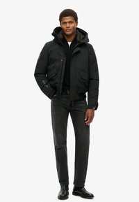 Sort dunjakke med lynlås foran, to forlommer og hætte. Båret over en sort sweater, parret med mørke jeans og sorte støvler.