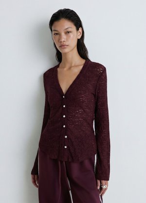 Donna che indossa un top bordeaux trasparente a maniche lunghe con bottoni e pantaloni coordinati, in piedi davanti a uno sfondo grigio chiaro uniforme.