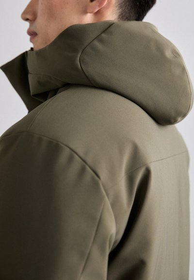Giubbotto imbottito verde oliva con un grande cappuccio strutturato e texture liscia. Presenta linee pulite e un design minimalista senza hardware visibile.