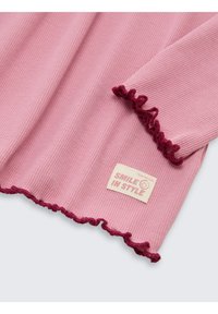 Roze geribbelde top met een rimpelige kastanjebruine zoom en een label met "SMILE IN STYLE." Zachte textuur met zichtbare horizontale lijnen.